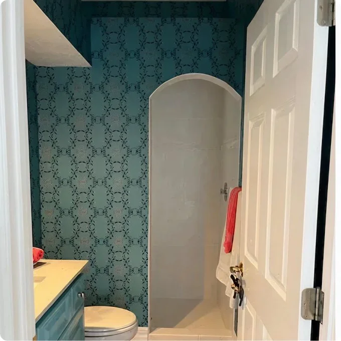 Small bathroom with patterned teal wallpaper, a compact shower stall with a curved door, a white door with a hook holding a white and red towel, and a partial view of a yellow sink or vanity.