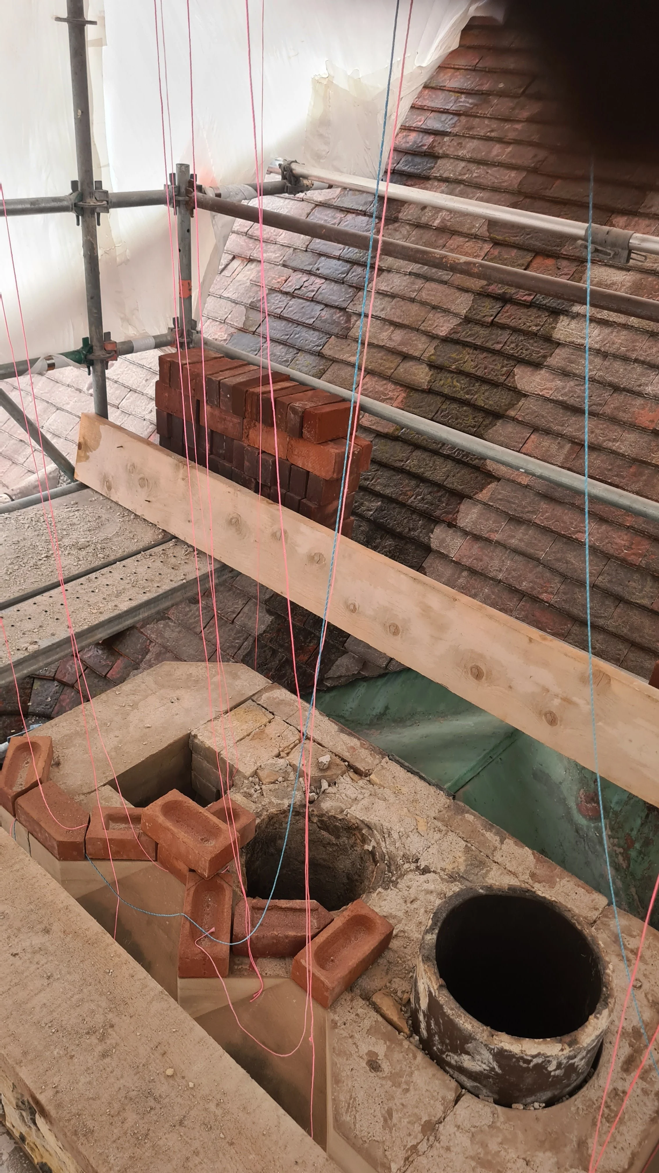 Construction site with scaffolding, bricks, and a construction hole, possibly for a chimney or pipe, on a roof with tiles and temporary protective covering.