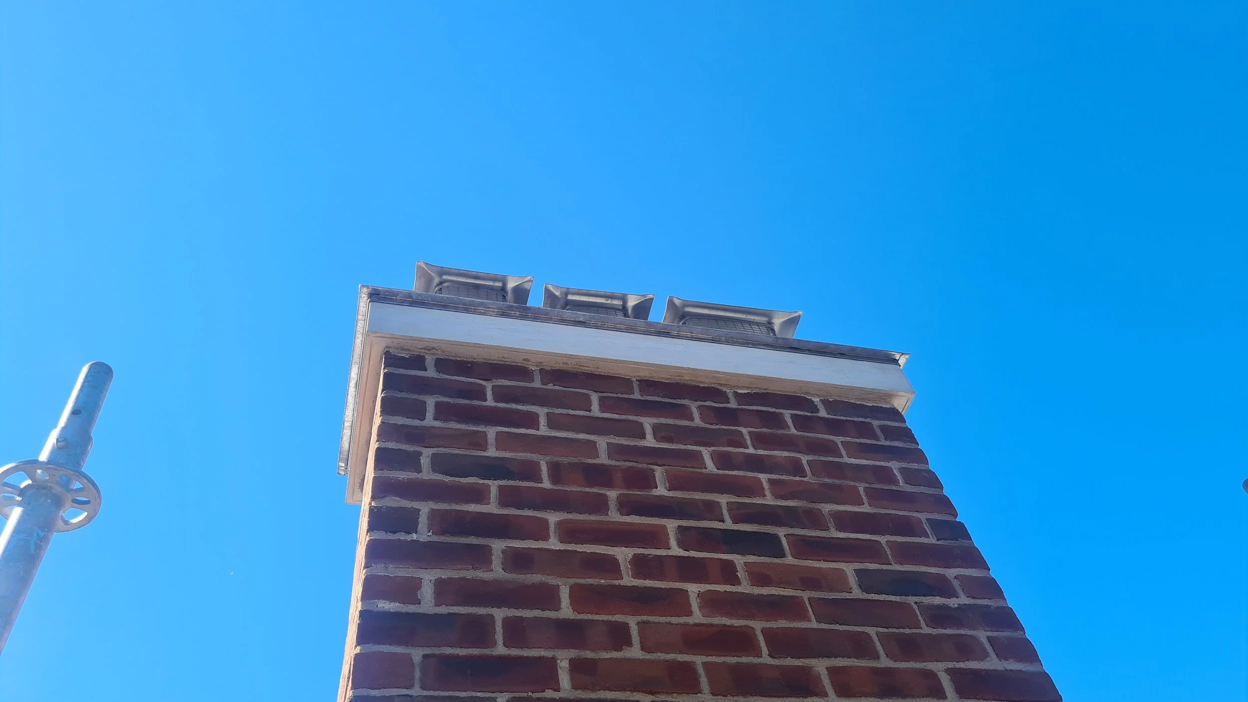 Close-up of a brick chimney with metal caps on top, against a clear blue sky. Part of a metal pipe is visible on the left side.
