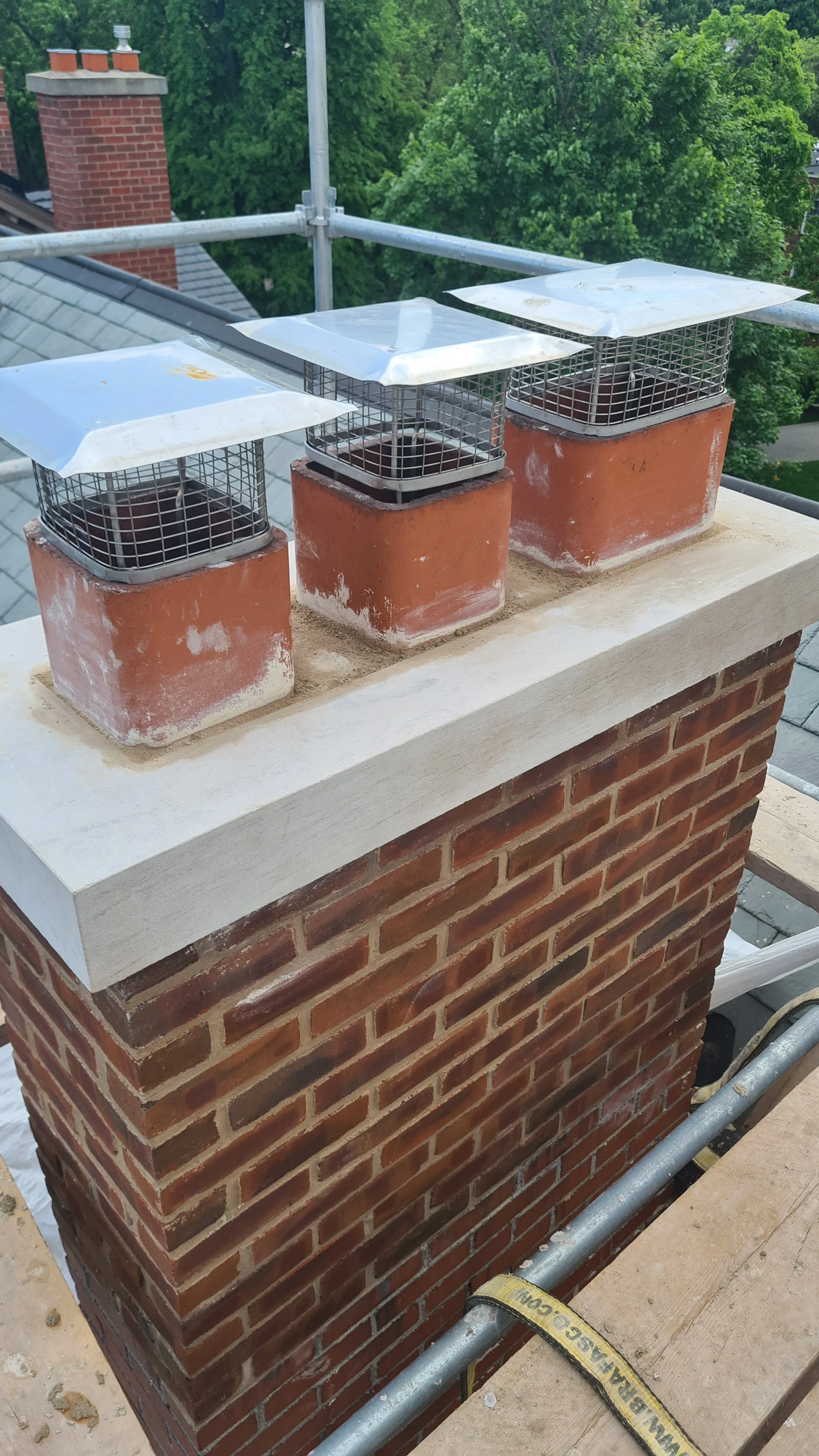 Three metal wire cages with metal covers on red brick chimneys, with a lush green tree and rooftops in the background.