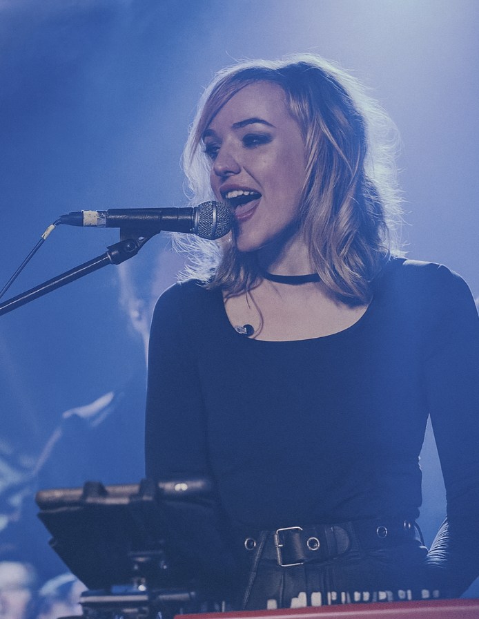 Young woman with blonde, wavy hair singing into a microphone on stage, wearing a black top and choker, with a keyboard in front of her.