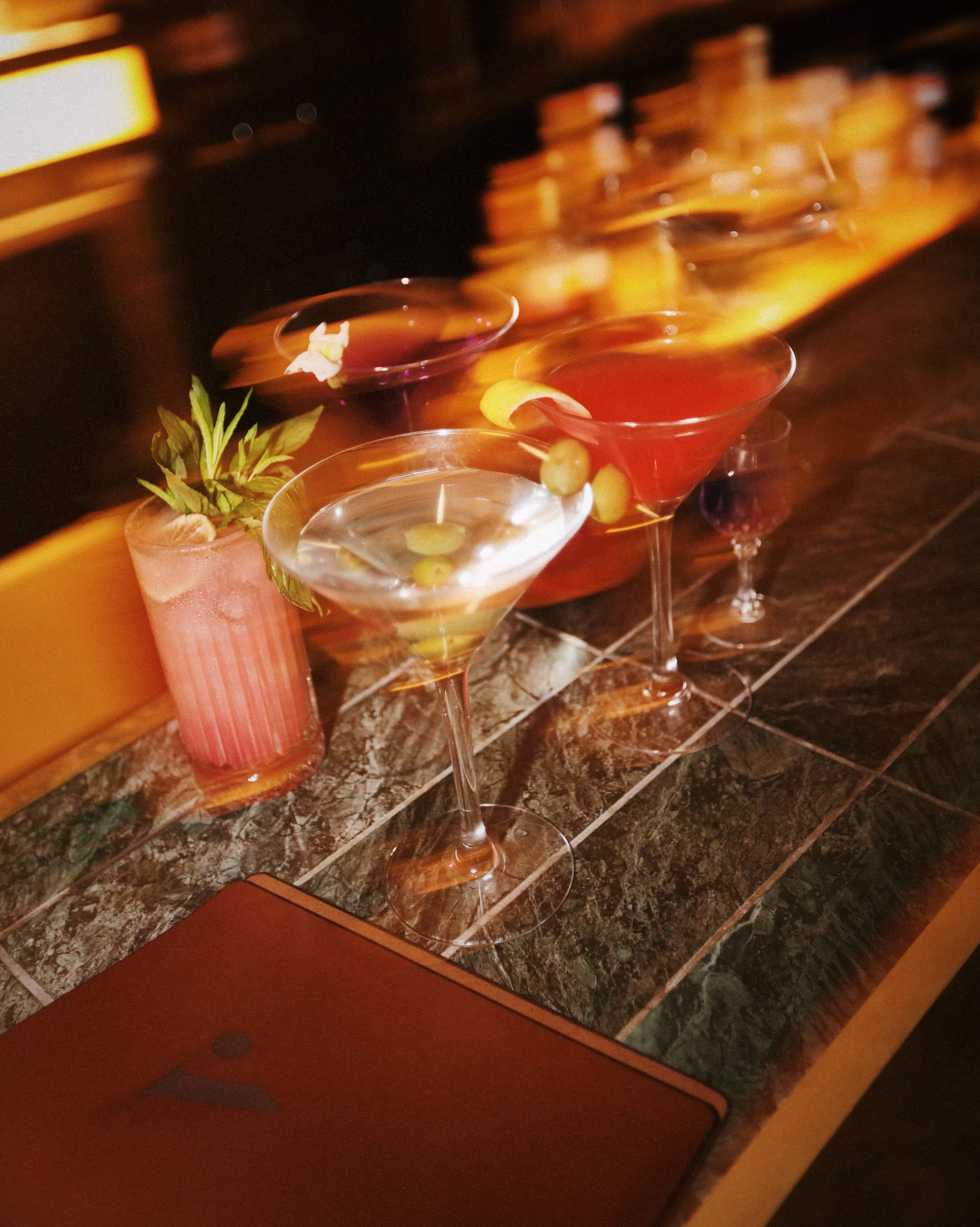 Four cocktails on a bar counter, with a pink drink garnished with mint, a clear drink with green grapes, a red drink, and a dark drink, along with a pink flower arrangement.