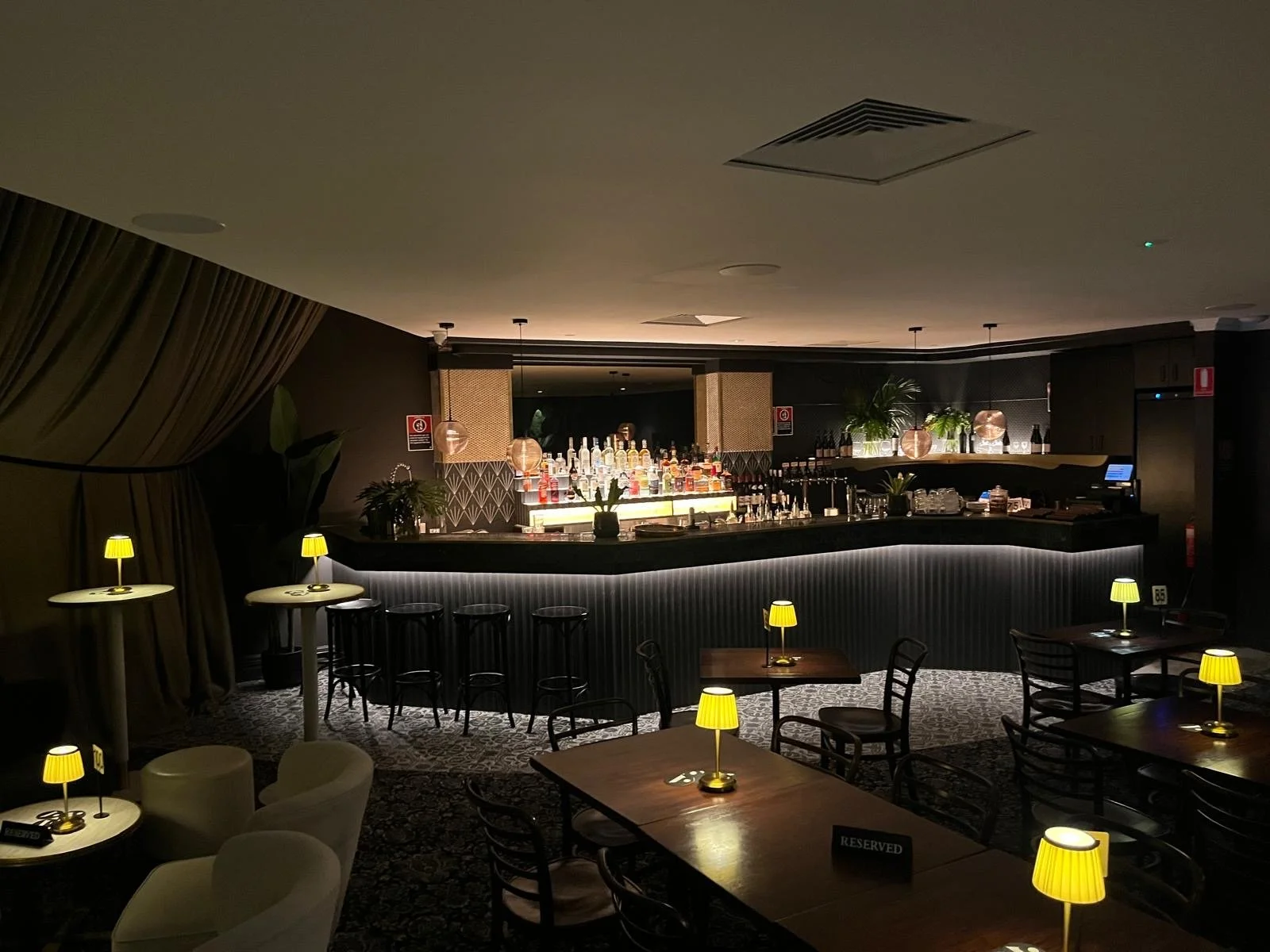 A dimly lit bar with a curved counter, bottles of liquor on shelves, small tables with yellow-lit lamps, chairs, and a reserved sign, with dark walls and plants.