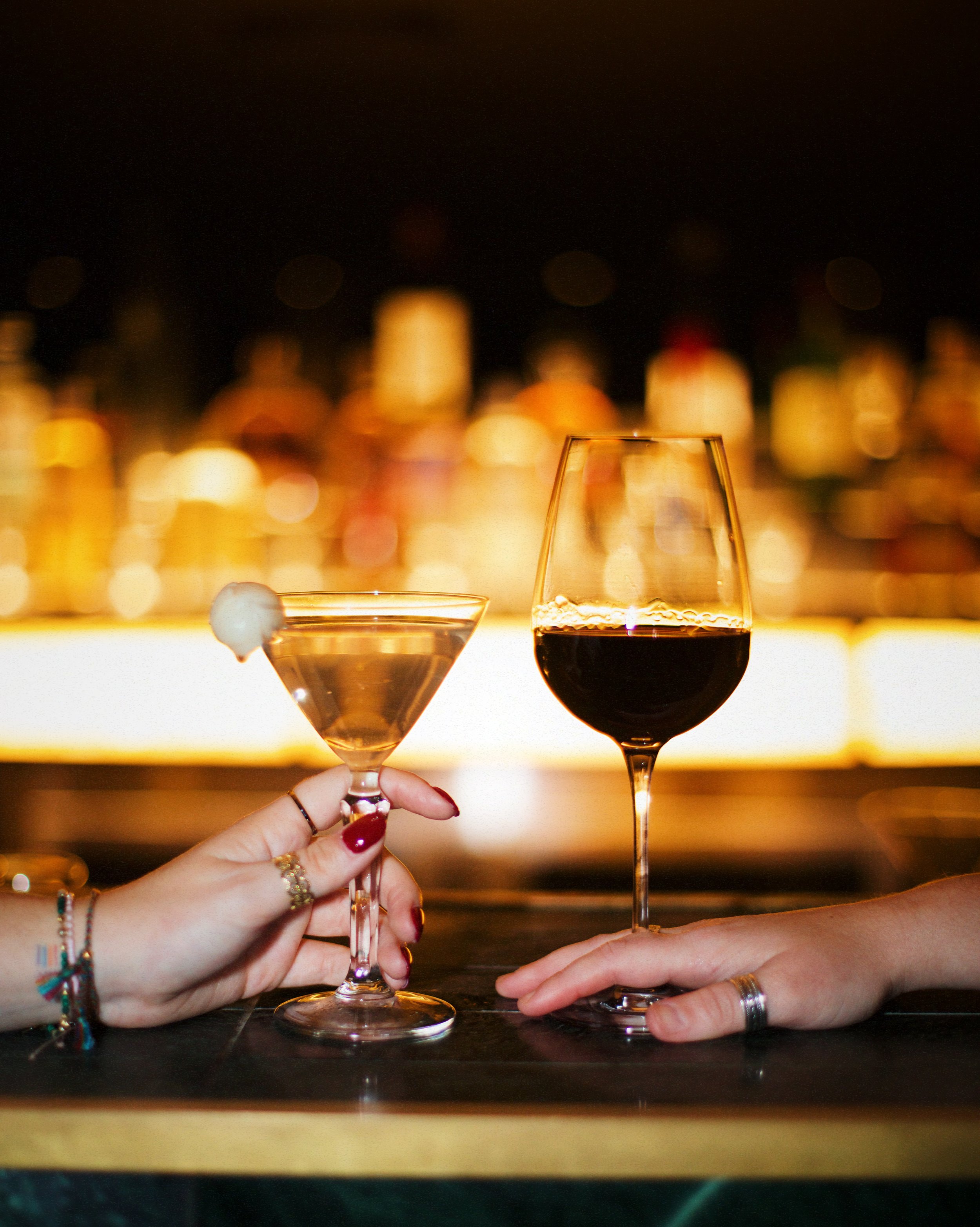 Two people toasting with a cocktail glass and a glass of red wine against a blurred cityscape at night.