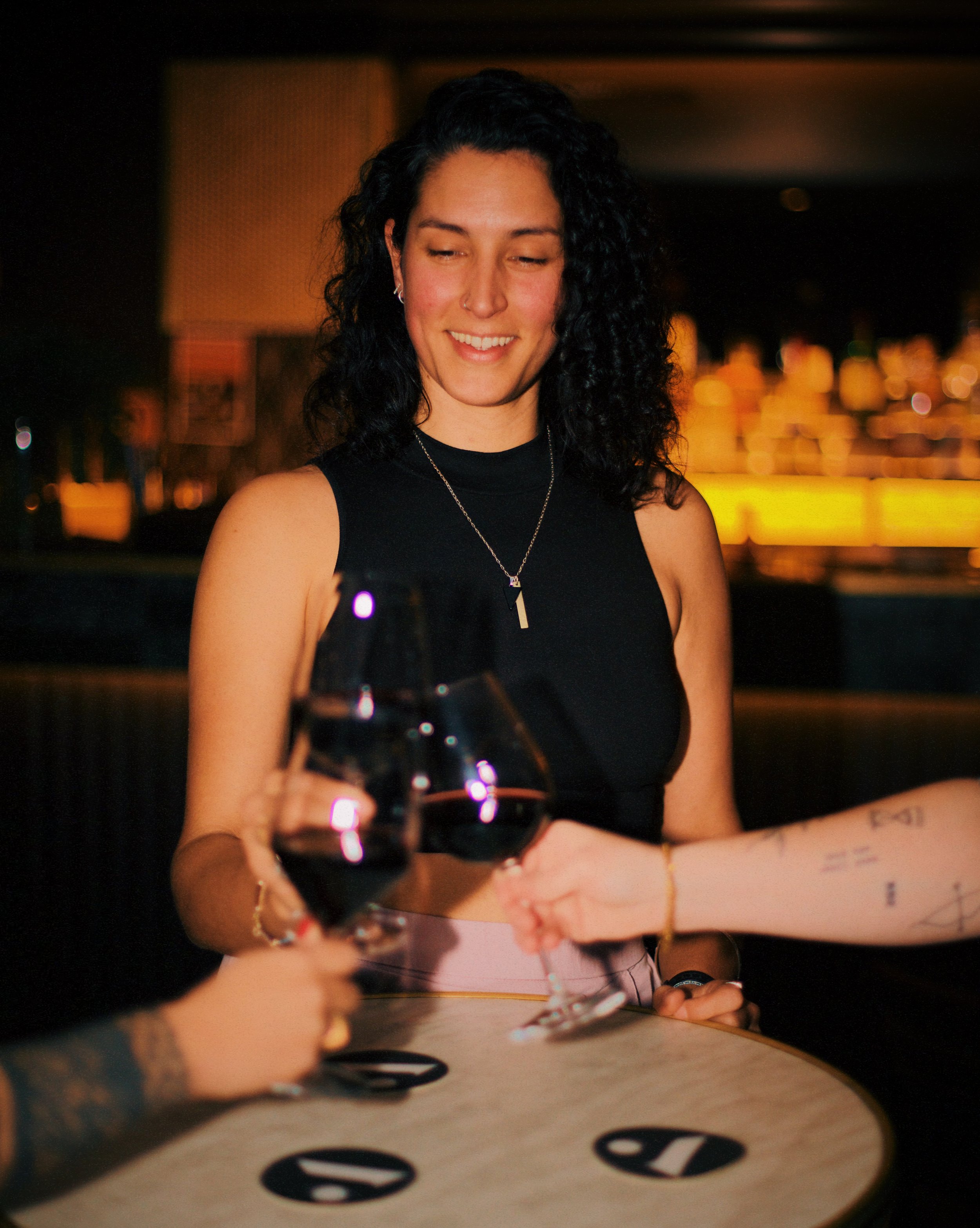 A woman with curly black hair in a black sleeveless top smiling and playing blackjack at a casino table with other players.