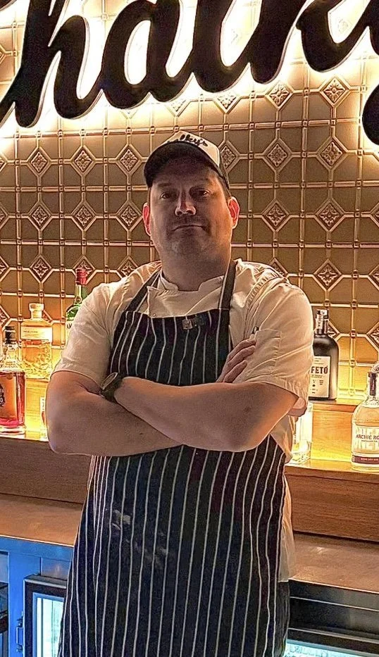 A chef standing with arms crossed in front of a bar, wearing a striped apron, white chef's coat, black cap, and watch, with various liquor bottles and a patterned tile wall behind him.