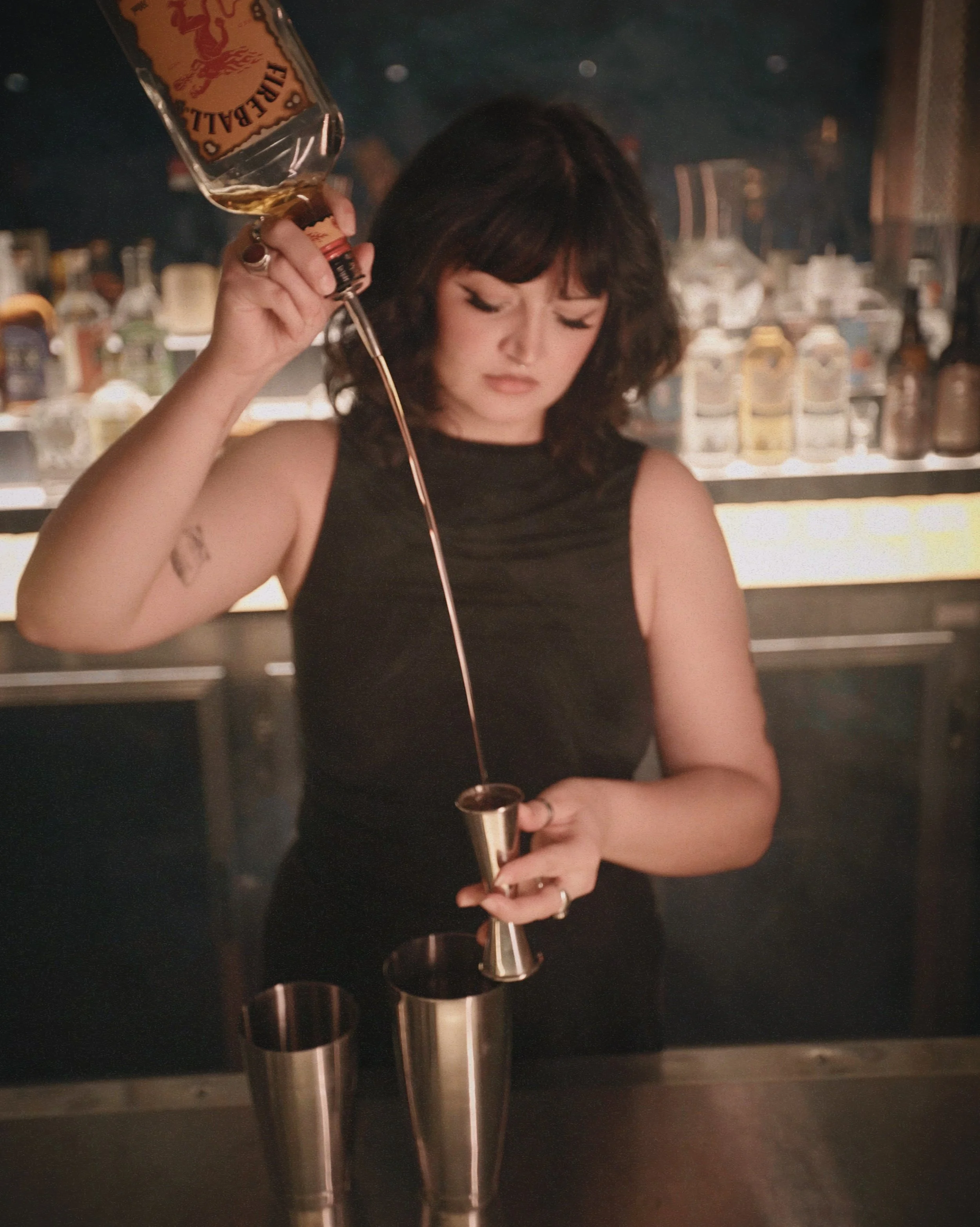 A woman with curly dark hair wearing a black sleeveless dress pouring a drink from a bottle into a jigger, in a bar with a backlit shelf of liquor bottles.