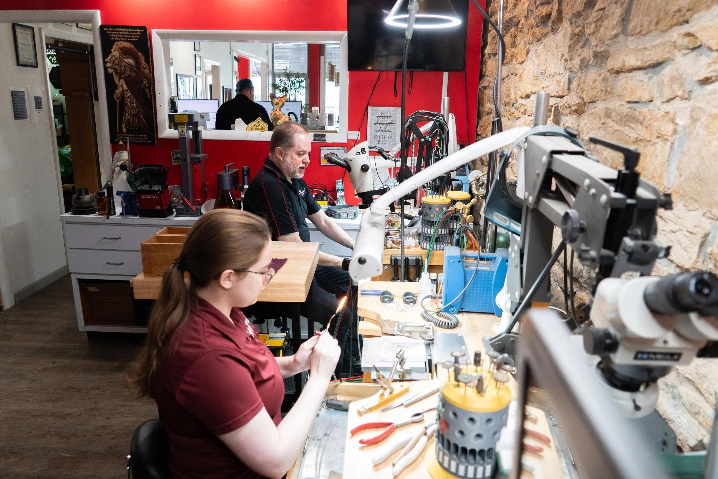 A woman working with wires and tools at a workbench, and a man using an electronic device at a different station, in a workshop with jewelry repair equipment and a stone wall.