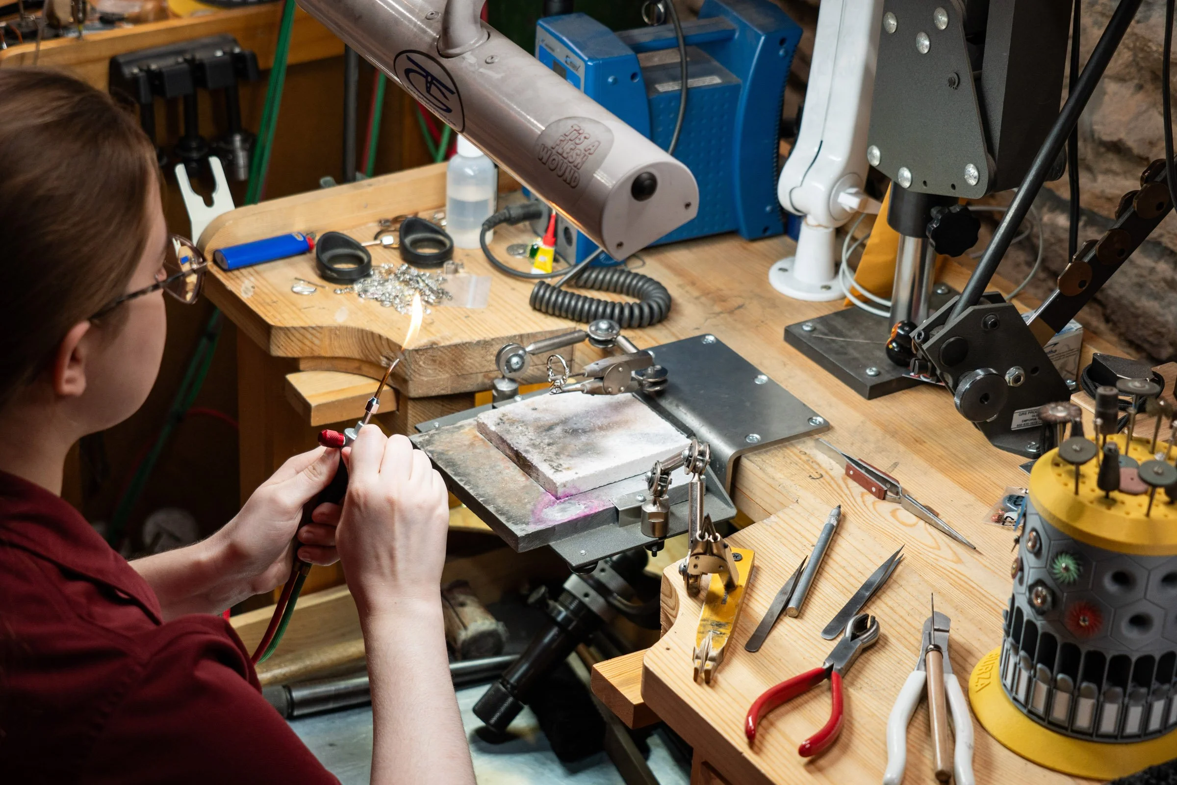 A person working at a jewelry-making workstation, soldering jewelry with various tools and equipment around.
