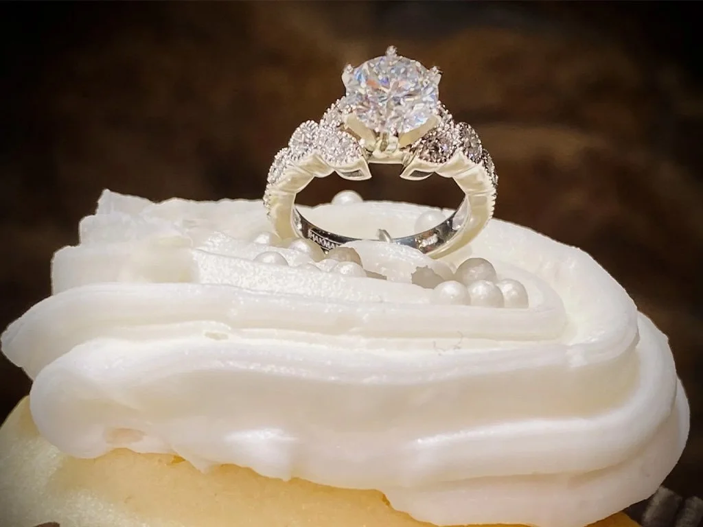 Close-up of an engagement ring with a large diamond on a cupcake topped with white frosting and pearls.