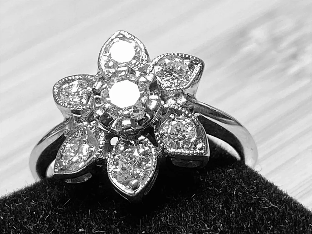 A silver ring with a large flower-shaped cluster of diamonds, resting on a black fabric surface.