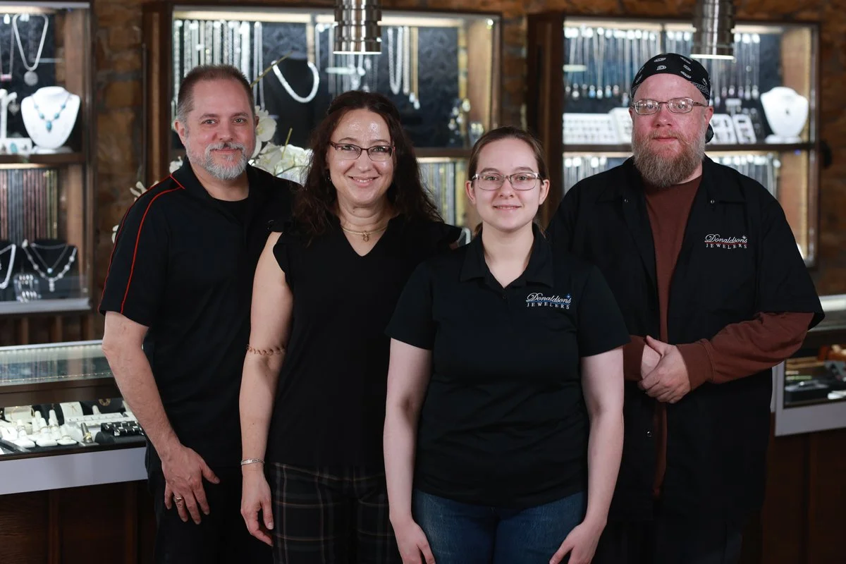 Four people standing inside a jewelry store, with display cases of jewelry behind them, smiling at the camera.