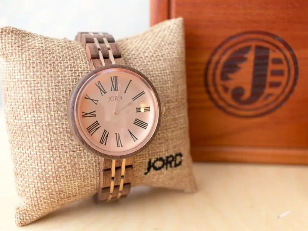 A James JORD wristwatch with a round face, Roman numerals, and a metal band, placed on a beige cushion with the JORD brand logo, and a wooden box with the JORD logo in the background.