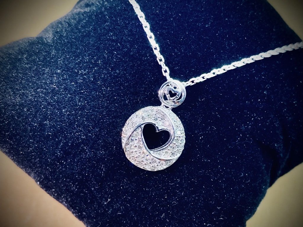 Silver necklace with a circular pendant containing a heart shape, adorned with small gemstones, displayed on a dark fabric background.