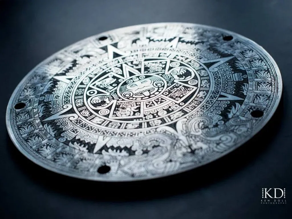 A silver round disc with intricate Aztec-style patterns and symbols, featuring a face at the center and surrounded by detailed geometric designs.