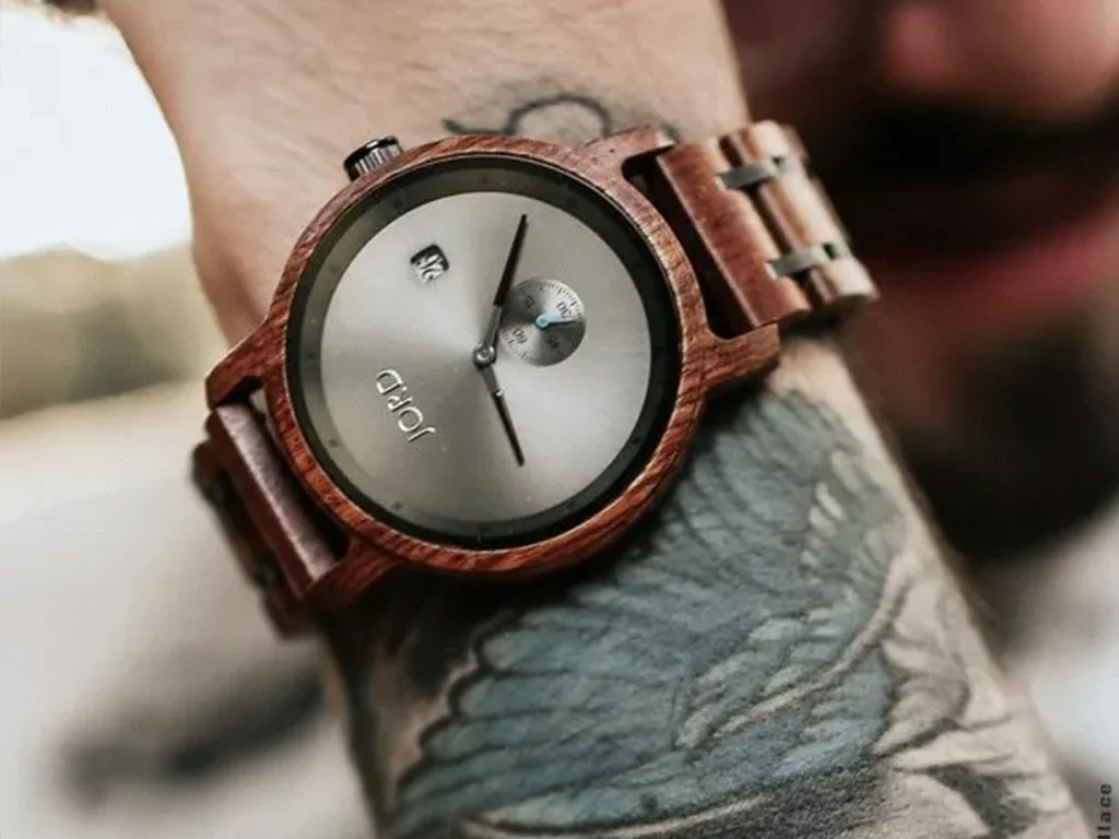 A wristwatch with a wooden band and a silver face showing the time and date, worn on a tattooed forearm.