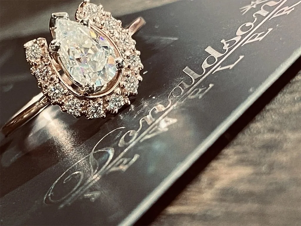 Close-up of a diamond engagement ring with a pear-shaped center stone surrounded by smaller diamonds on a black surface with embossed silver lettering.