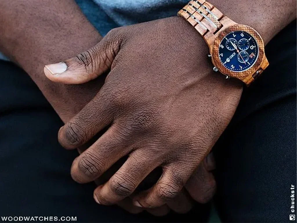 Close-up of a man's dark-skinned hands resting on his lap, wearing a wooden wristwatch with a blue dial.