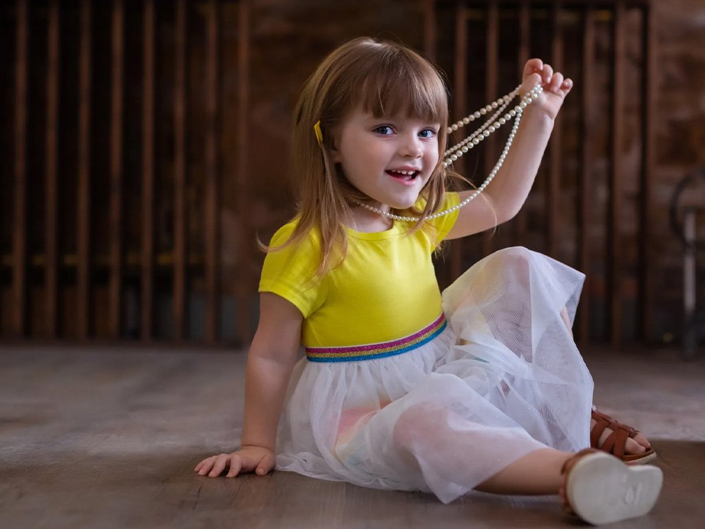 A young girl sitting on the wooden floor, holding a string of pearls, wearing a bright yellow top and a white tulle skirt, with a brown sandal on one foot, smiling at the camera.