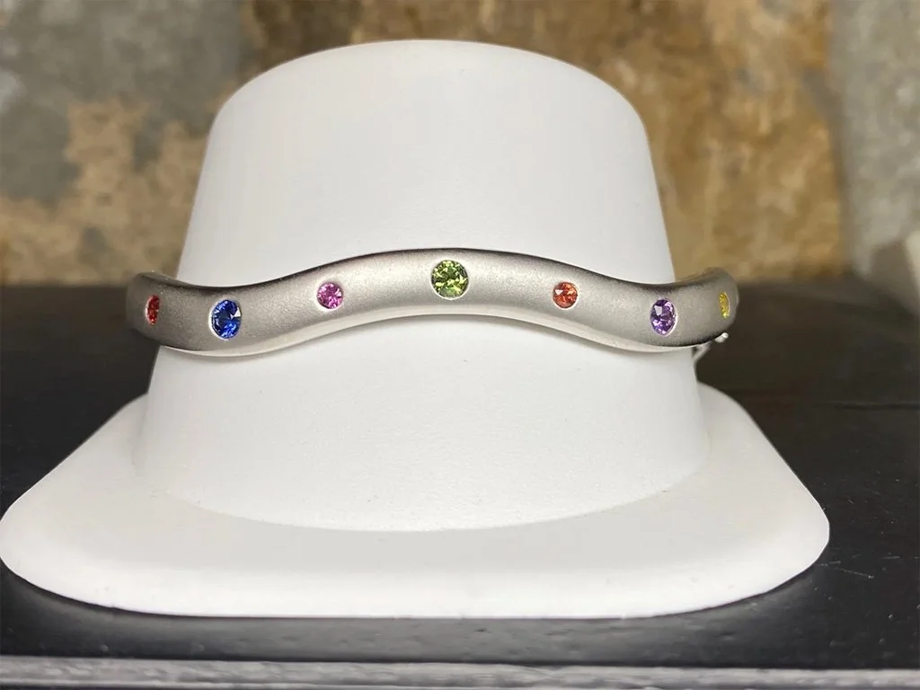 A silver bracelet with colorful gemstones evenly spaced around it, displayed on a white stand.