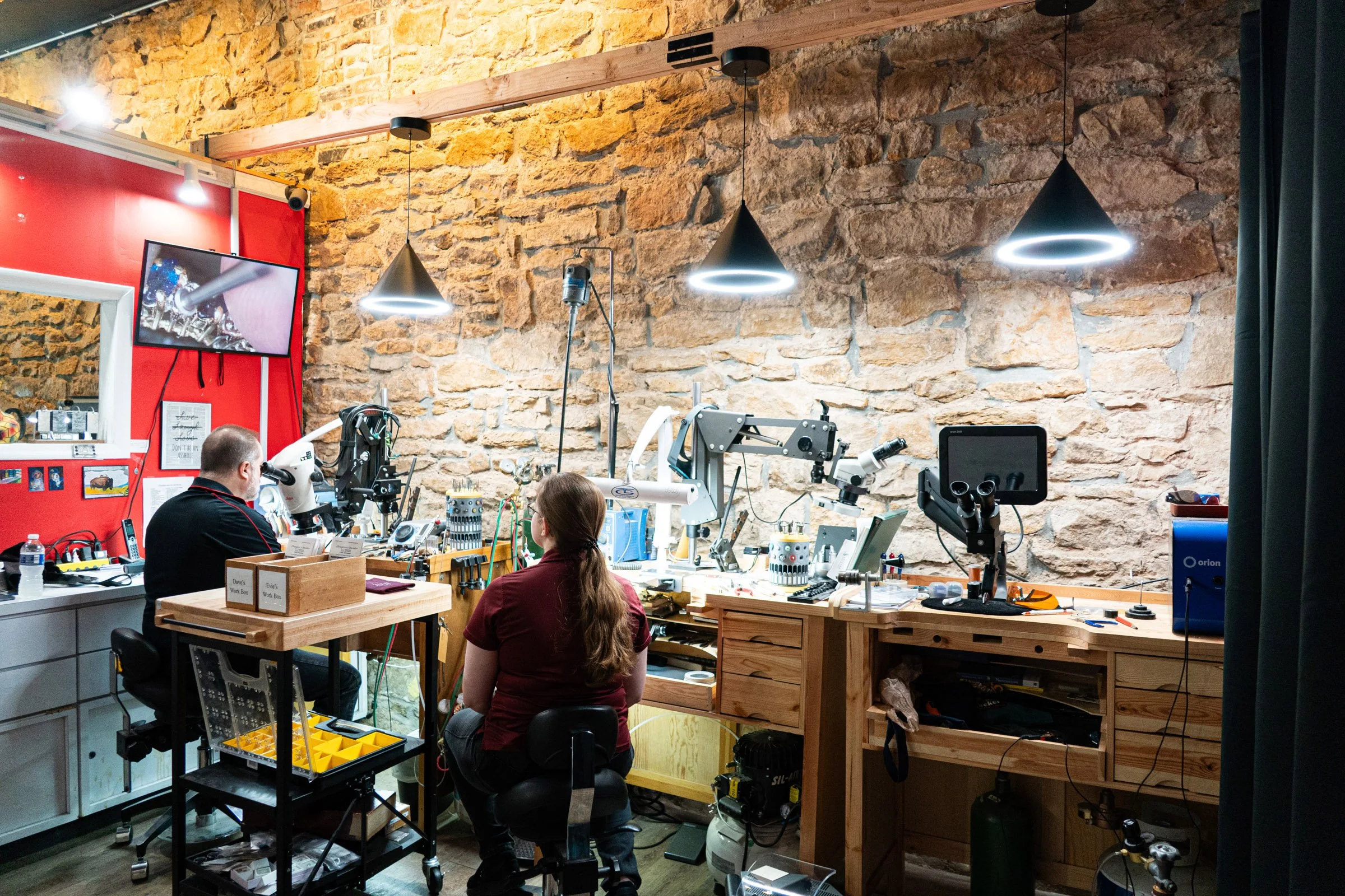 A jewelry repair shop with two people working on jewelry at a wooden workbench against a brick wall in Topeka Kansas.