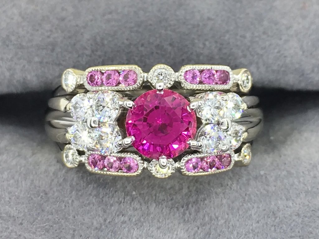 A silver ring featuring a central pink gemstone, surrounded by two diamonds, with additional pink gemstones and diamonds set in the band.