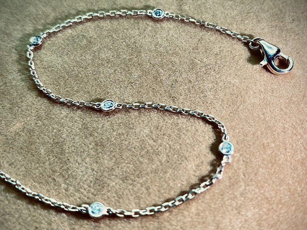 Silver chain necklace with small round clear gemstones evenly spaced along it, laid out on a textured brown surface.