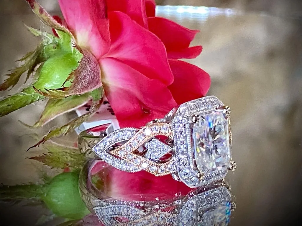 A close-up of an ornate diamond engagement ring with a large center stone, positioned on a reflective surface next to a pink flower with green leaves.