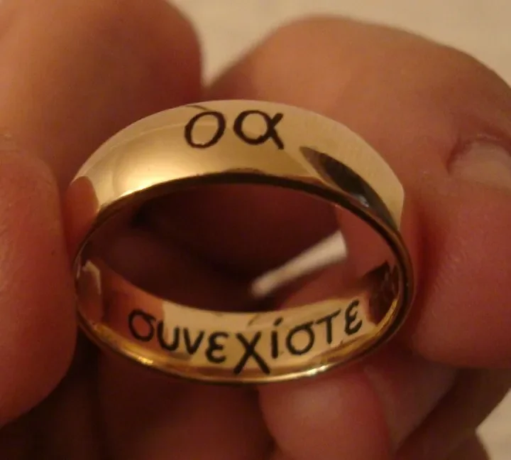 A person holding a silver ring with engraved Greek text. The ring reflects part of the person's fingers and the surrounding environment.