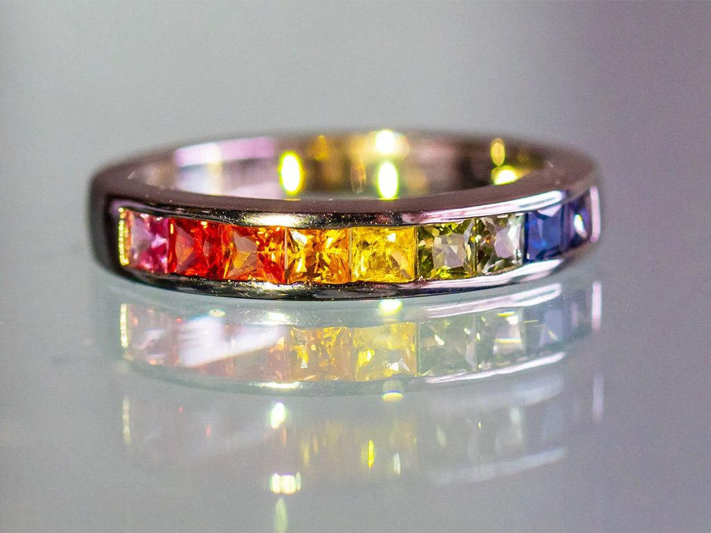 A silver ring with square-cut gemstones in rainbow colors, arranged in a gradient from red to purple.