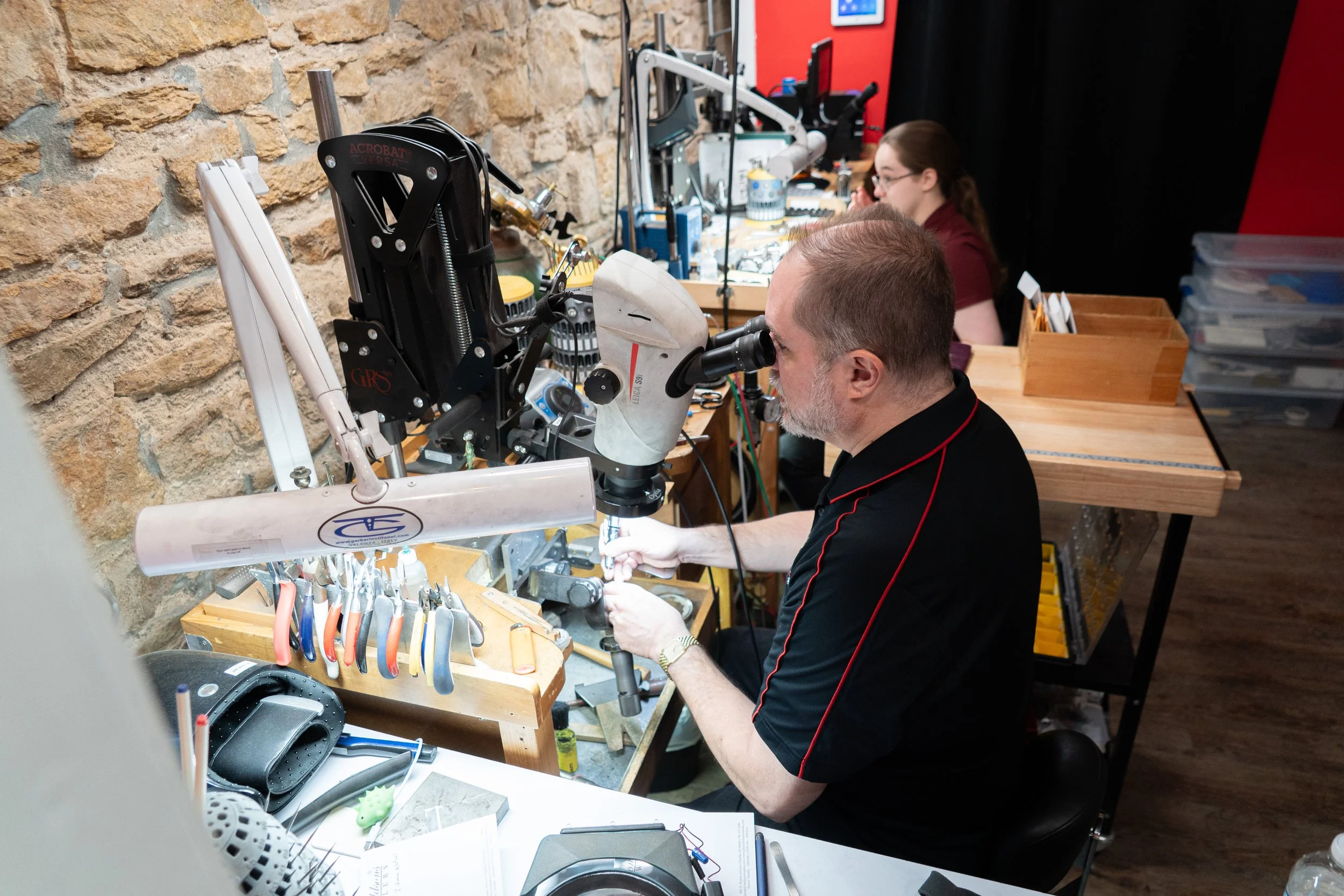 A man working at a jewelry repair shop in Topeka Kansas with a microscope, surrounded by tools and equipment, with a woman working in the background.