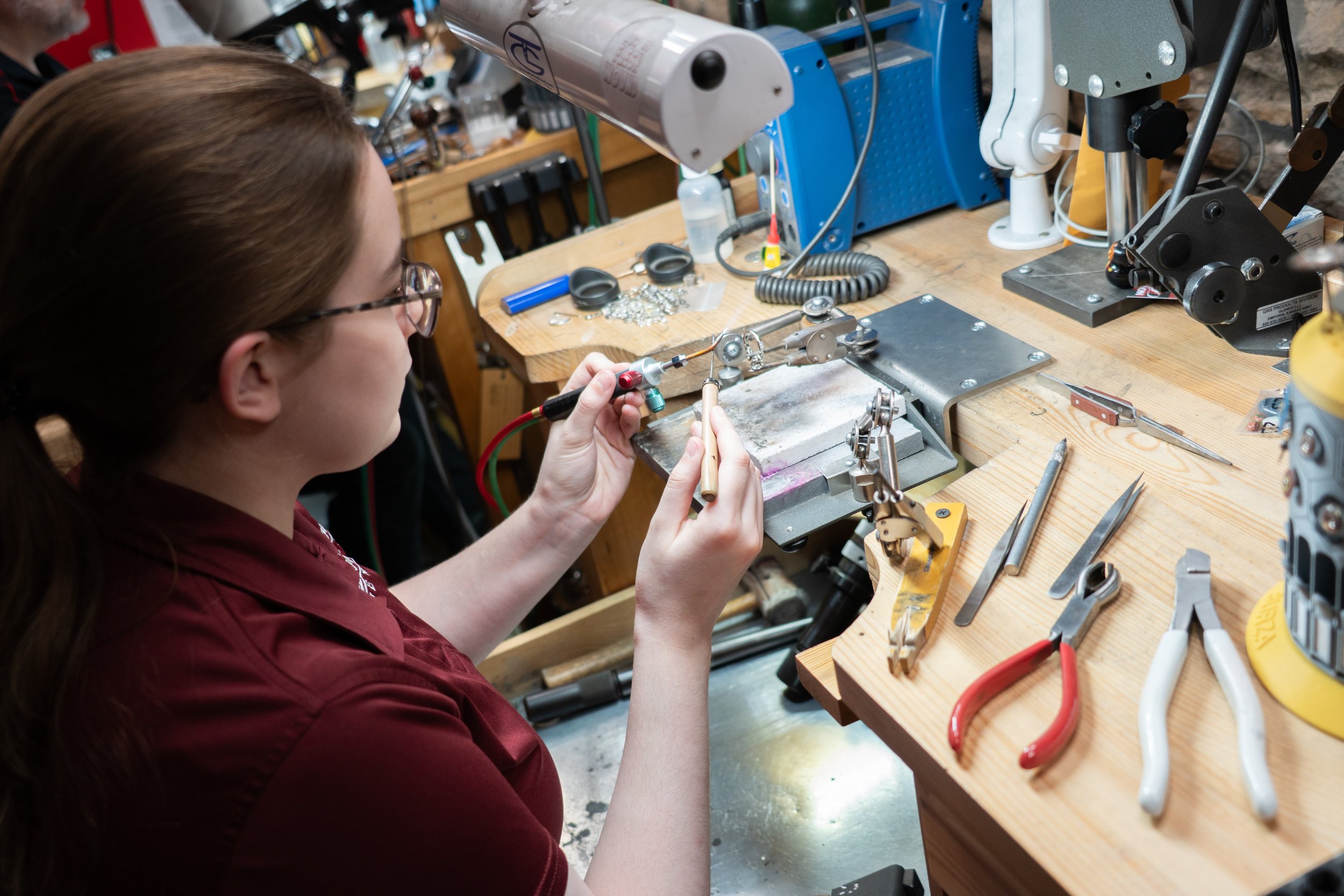 A woman working at a jewelry repair station in Topeka with various tools on a wooden workbench, including pliers, screwdrivers, and a soldering iron.