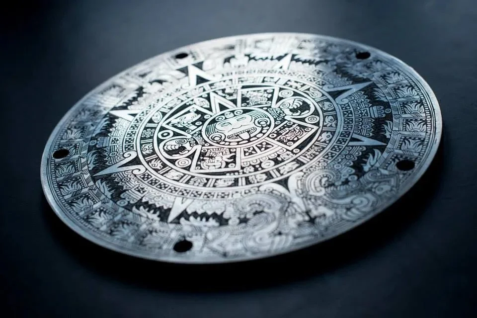 A round, black and white engraved disc with intricate patterns and designs inspired by Mayan or Aztec calendar art, featuring a central face and various symbolic elements.