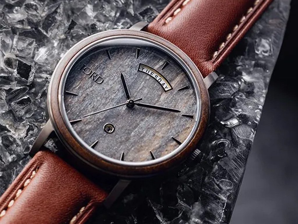 Close-up of a wristwatch with a wooden bezel, gray textured dial, and brown leather strap. The watch shows the day as Tuesday and the date as the 4th. The background is a bed of ice.