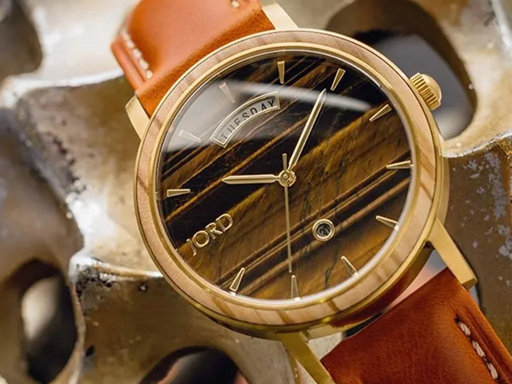 Close-up of a JORD watch with a wood grain dial and gold accents, leather strap, and day/date display, resting on a textured metallic surface.