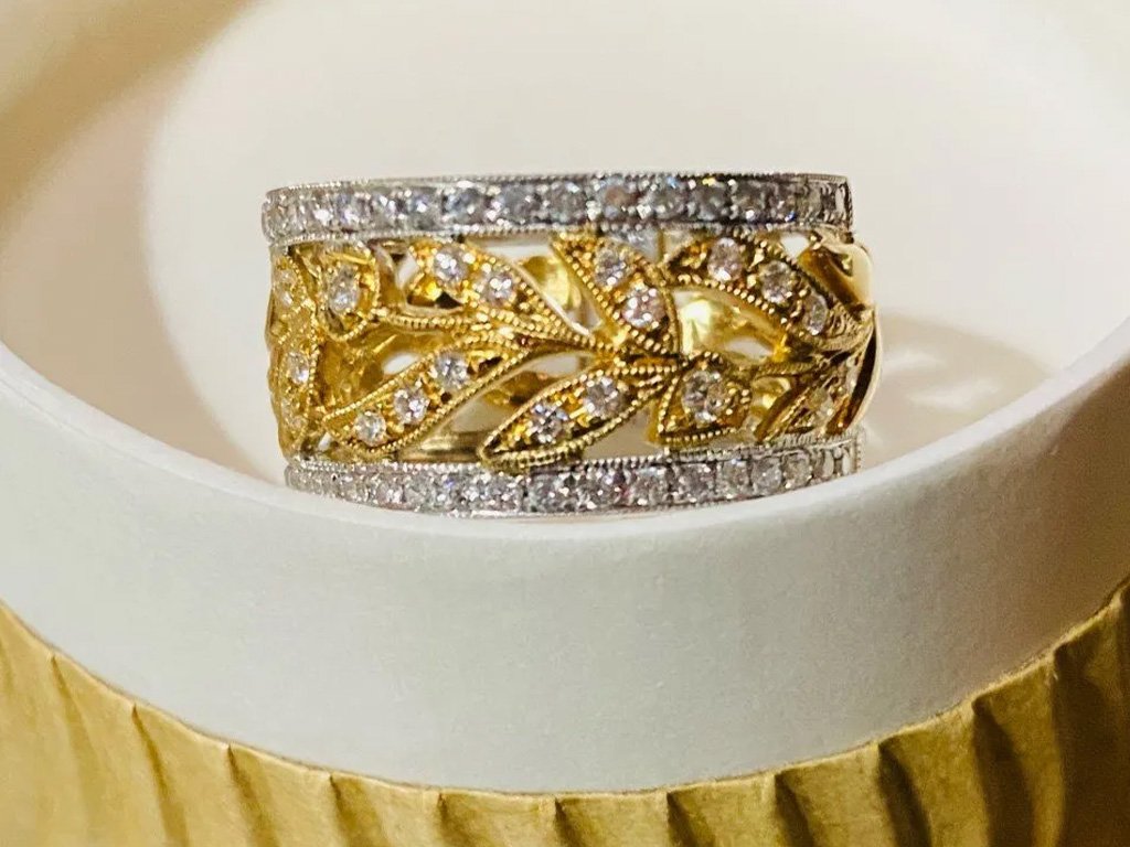 A close-up of a gold and silver ring with intricate leaf and vine design, encrusted with small diamonds, placed inside a round white jewelry box.