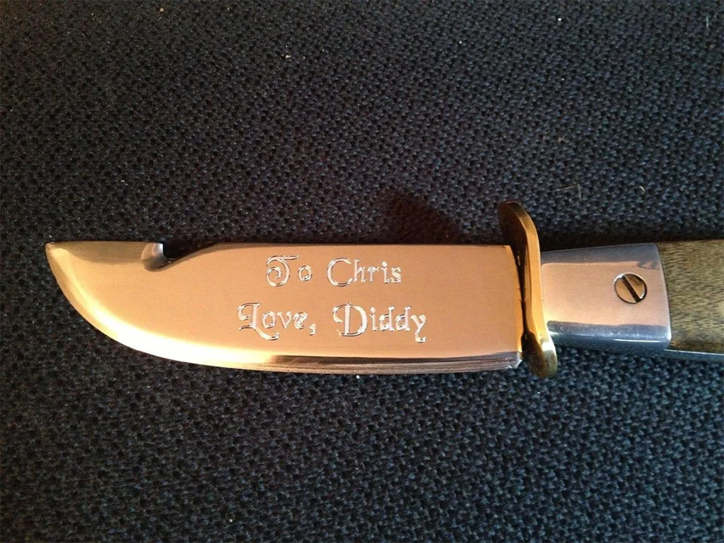 A pocket knife with a silver blade engraved with the words 'To Chris, Love, Diddy' on a black textured surface.