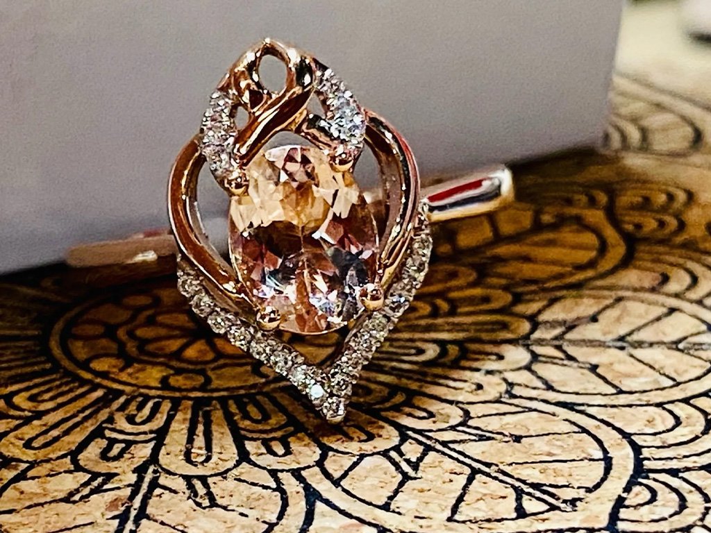 A close-up of a gold ring with a large oval gemstone surrounded by smaller stones, placed on a wooden surface with an intricate pattern.