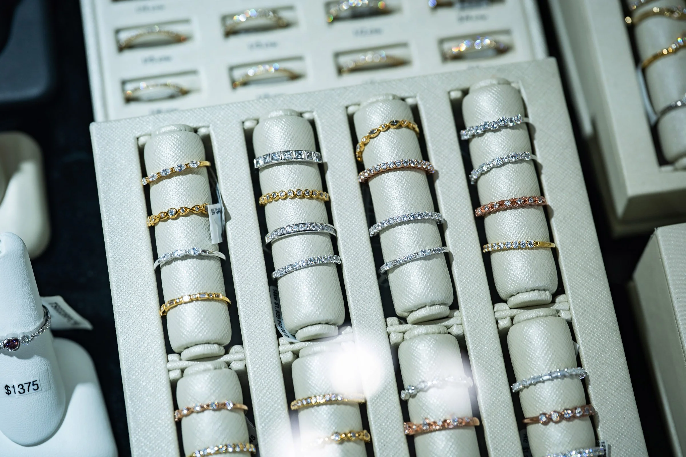 Display of various rings in a jewelry store, arranged on white rings holders with multiple gold and silver rings encrusted with diamonds.