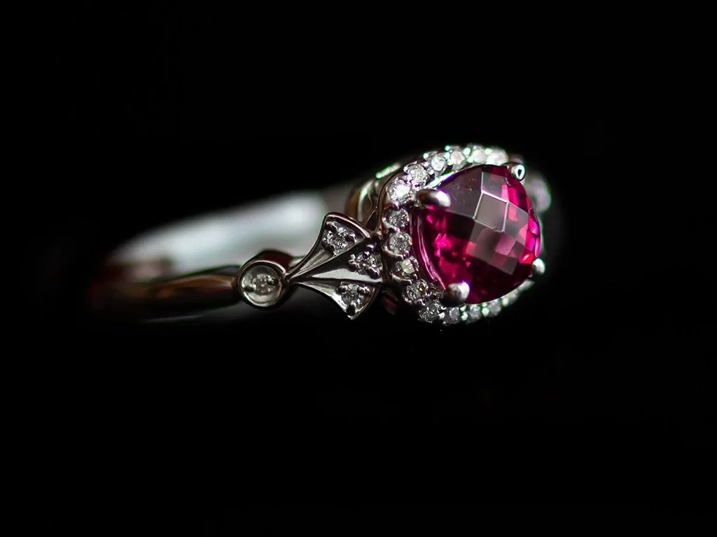 Silver ring with a large faceted red gemstone and small diamonds surrounding it on a black background.