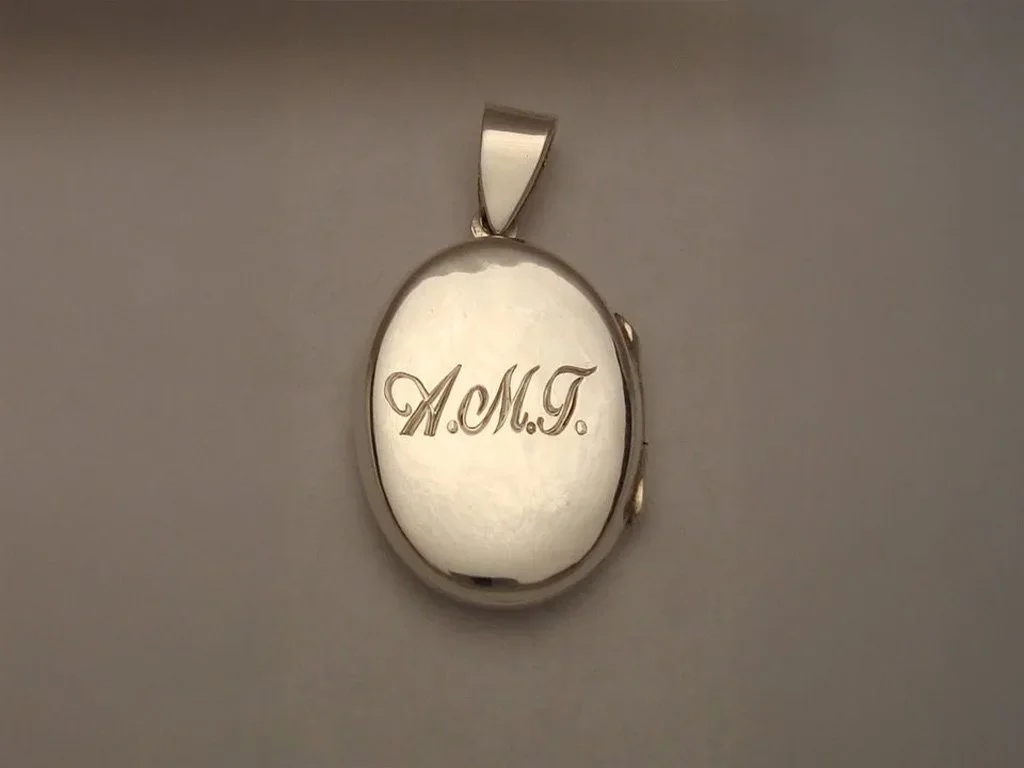 A silver oval locket with the initials 'A.M.T.' engraved on it, hanging on a plain wall.