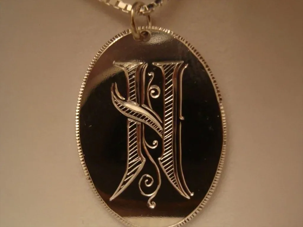 Silver oval pendant with an ornate, stylized letter 'N' engraved on a black background, attached to a chain.