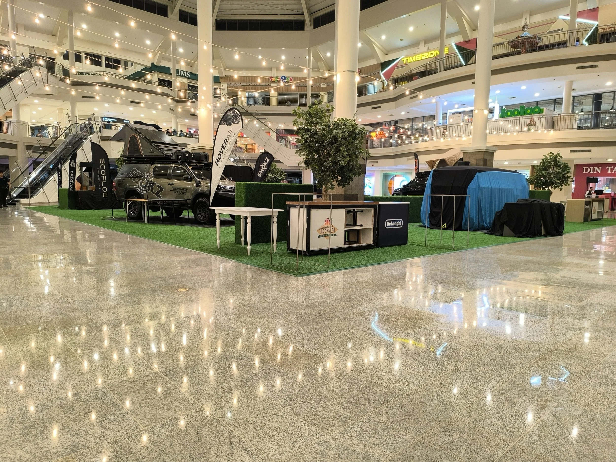Interior of a multi-level shopping mall with escalators and bright lighting. There is an outdoor display of off-road vehicles, tents, and banners in the foreground, while store signs are visible on the upper floors.