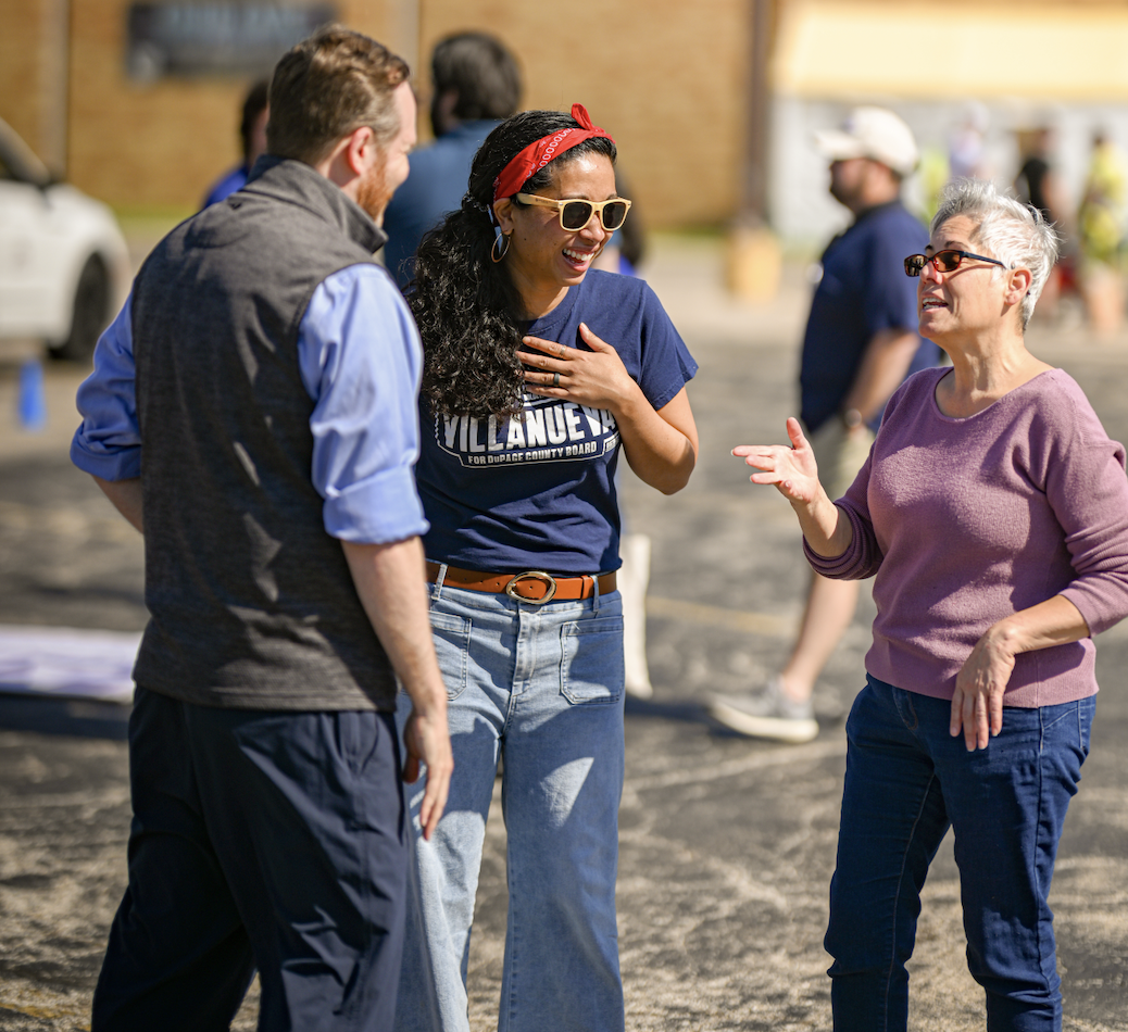 Three people are engaged in a lively conversation outdoors, smiling and gesturing. The woman in the middle is wearing sunglasses, a red headband, and a blue T-shirt, while the woman on the right is dressed in a pink sweater and dark jeans. The man on the left is wearing a vest over a light blue shirt. They are at an outdoor event or gathering with other people and structures in the background.