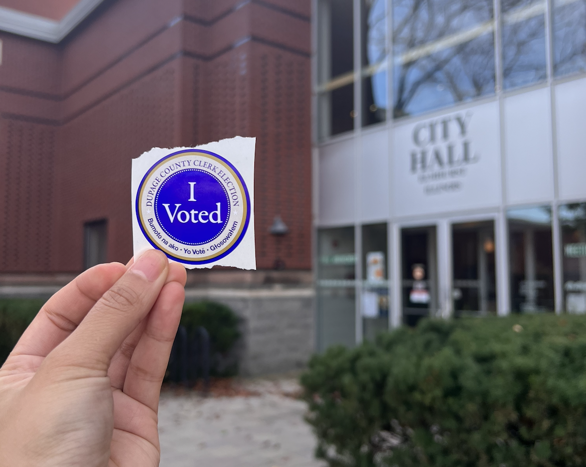 Close-up of a hand holding an 'I Voted' sticker in front of a city hall building.