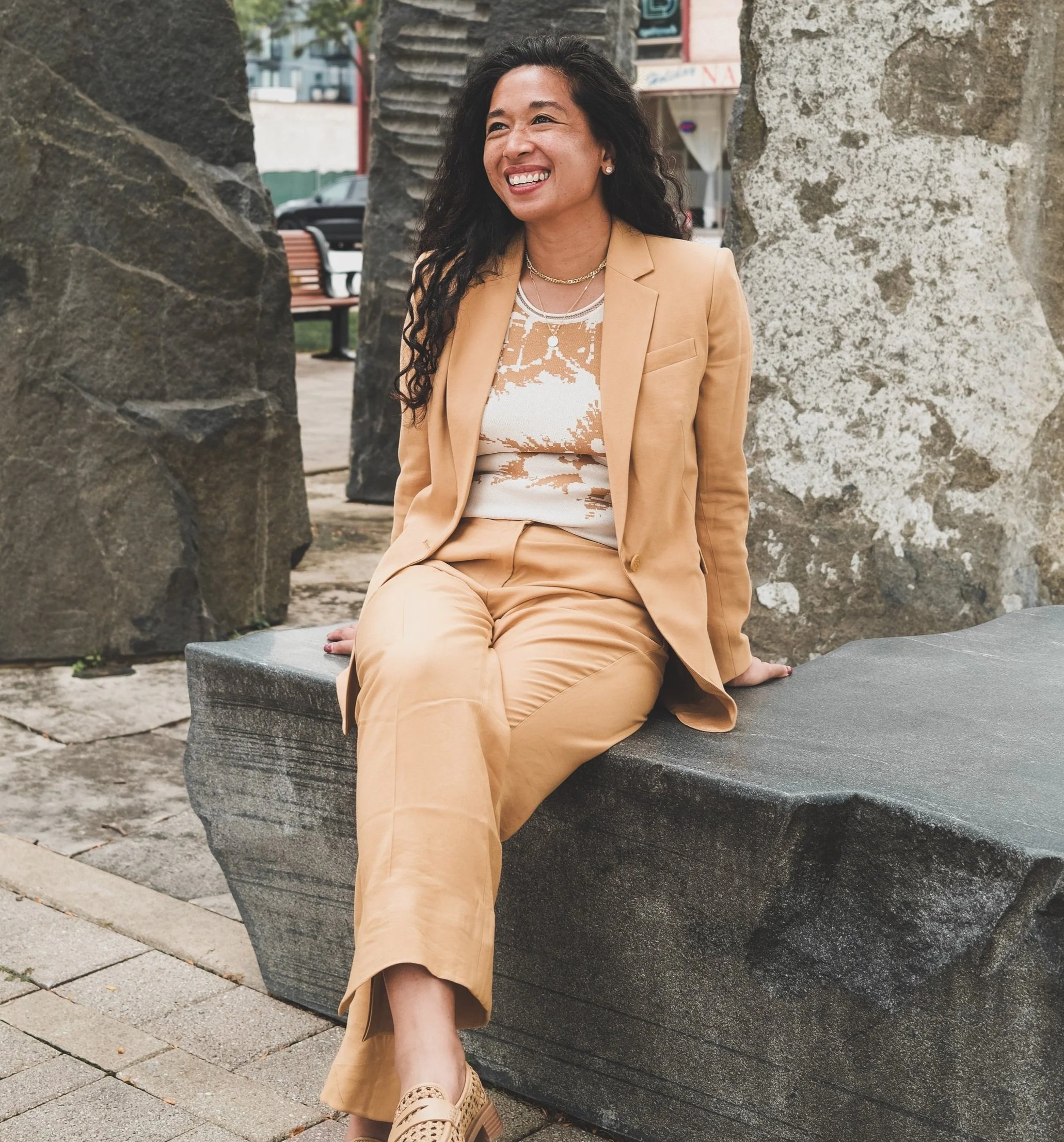 Woman sitting on a bench outdoors, smiling, wearing a tan suit and patterned shoes, with stone pillars in the background.