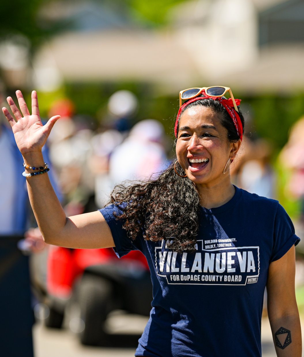 A woman smiling and waving, wearing a navy blue campaign shirt that says 'Villanueva for DuPage County Board,' with sunglasses on her head, a red bandana, and bracelets, outdoors on a sunny day with other people blurred in the background.
