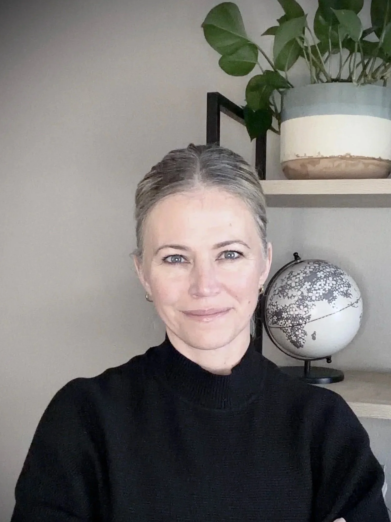 A woman with hair pulled back and blue eyes smiling at the camera, wearing a black sweater. Behind her is a bookshelf with a potted plant and a globe.
