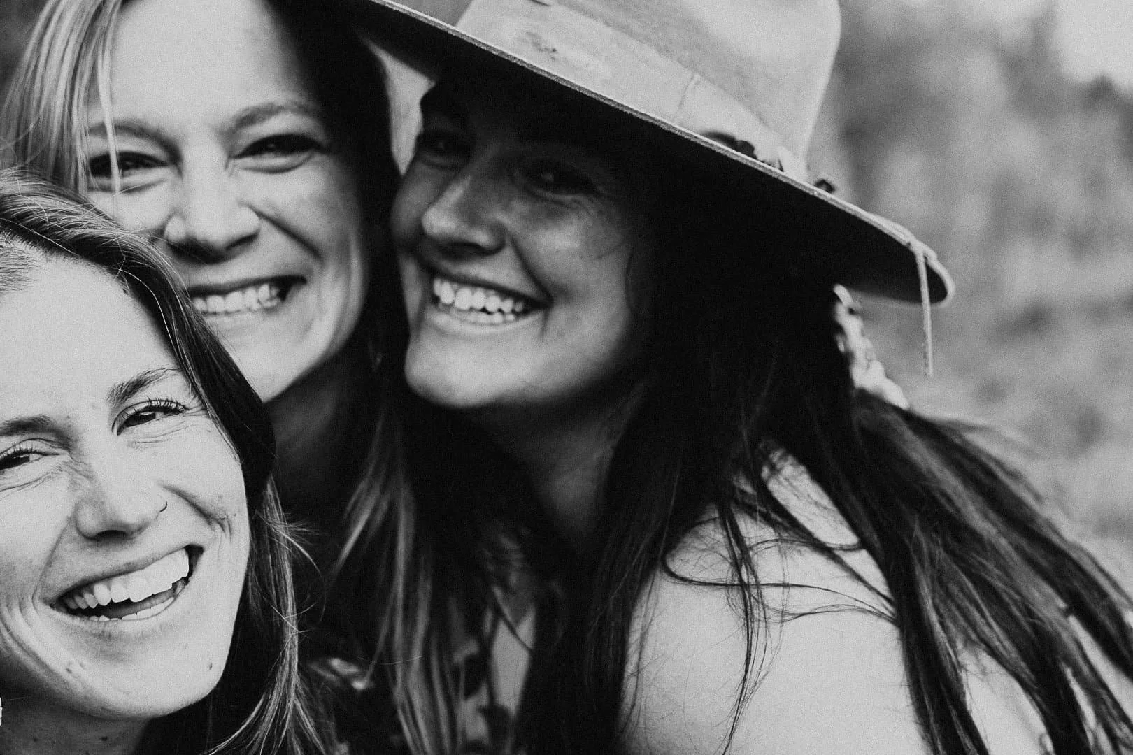 A black-and-white close-up photo of three smiling women leaning their heads together in a joyful moment. The woman on the right is wearing a wide-brimmed hat.