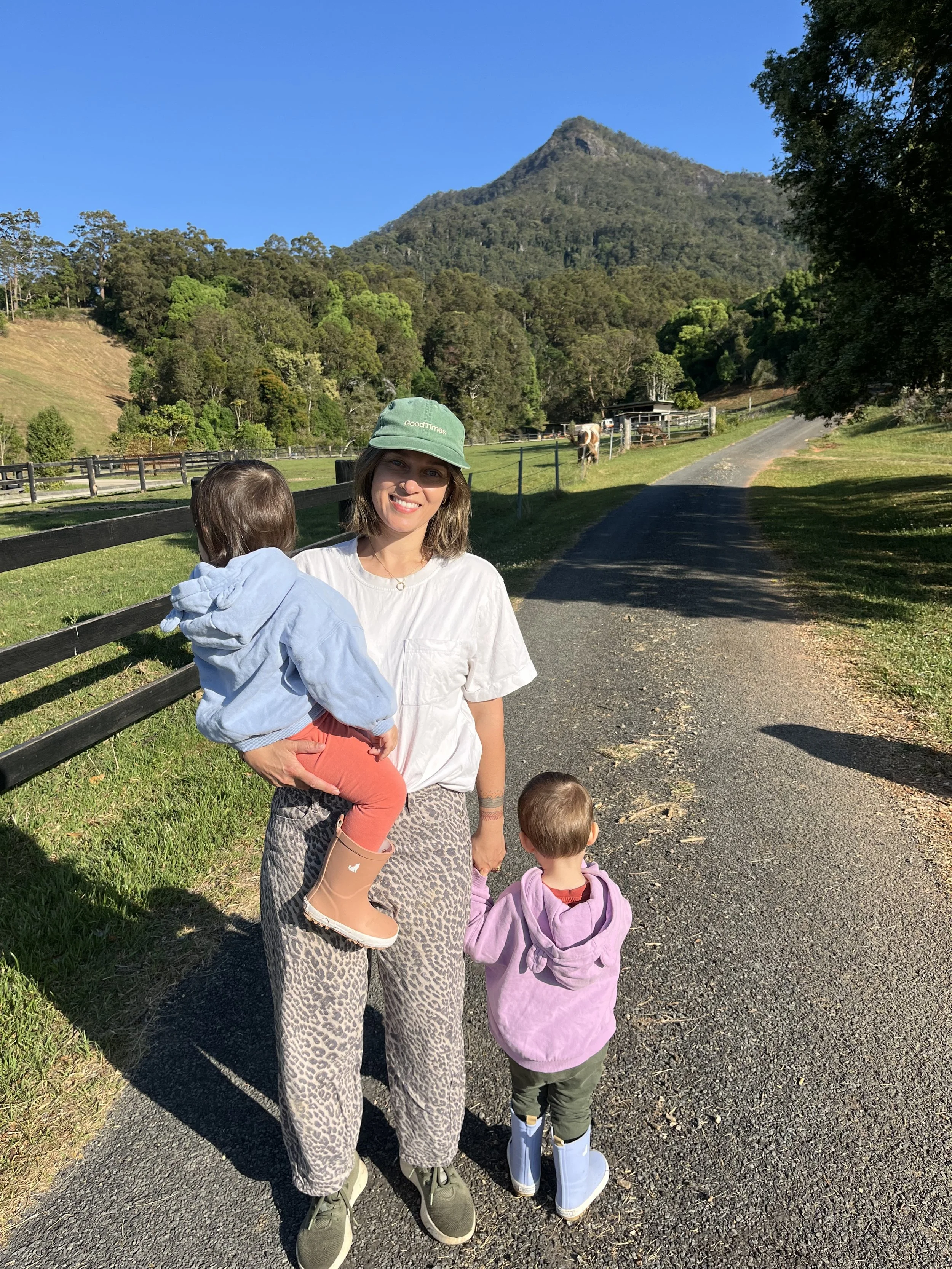 A woman with a hat smiling, holding a young girl on her hip, and a small boy standing beside her, outdoors on a gravel path with green grass, trees, a mountain, and a blue sky in the background.